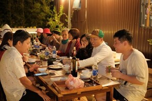 A group of people seated around a wooden table with various dishes and drinks, suggesting a communal dining setting. - Mountain Nest Villa Dalat - BBQ, Cloud Hunting, Camping (Lac Duong District)