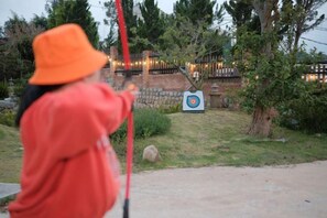 A person aiming a bow at a target in an outdoor setting with trees and a brick wall in the background. - Mountain Nest Villa Dalat - BBQ, Cloud Hunting, Camping (Lac Duong District)