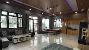 A spacious living area with a sofa, a coffee table, and a TV. There are large windows and a kitchen area visible. - Mountain Nest Villa Dalat - BBQ, Cloud Hunting, Camping (Lac Duong District)