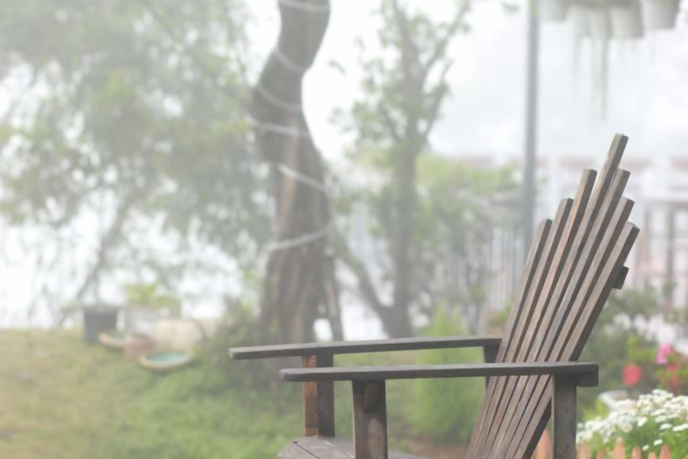 Una silla de madera con respaldo y reposabrazos, ubicada al aire libre con un fondo desenfocado de vegetación y un edificio.