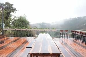 A wooden deck with a long table and chairs, overlooking a misty landscape. - Mountain Nest Villa Dalat - BBQ, Cloud Hunting, Camping (Lac Duong District)