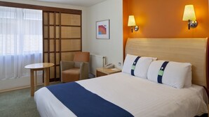 Habitación de hotel con cama, una silla, una mesita, una lámpara y un cuadro en la pared.