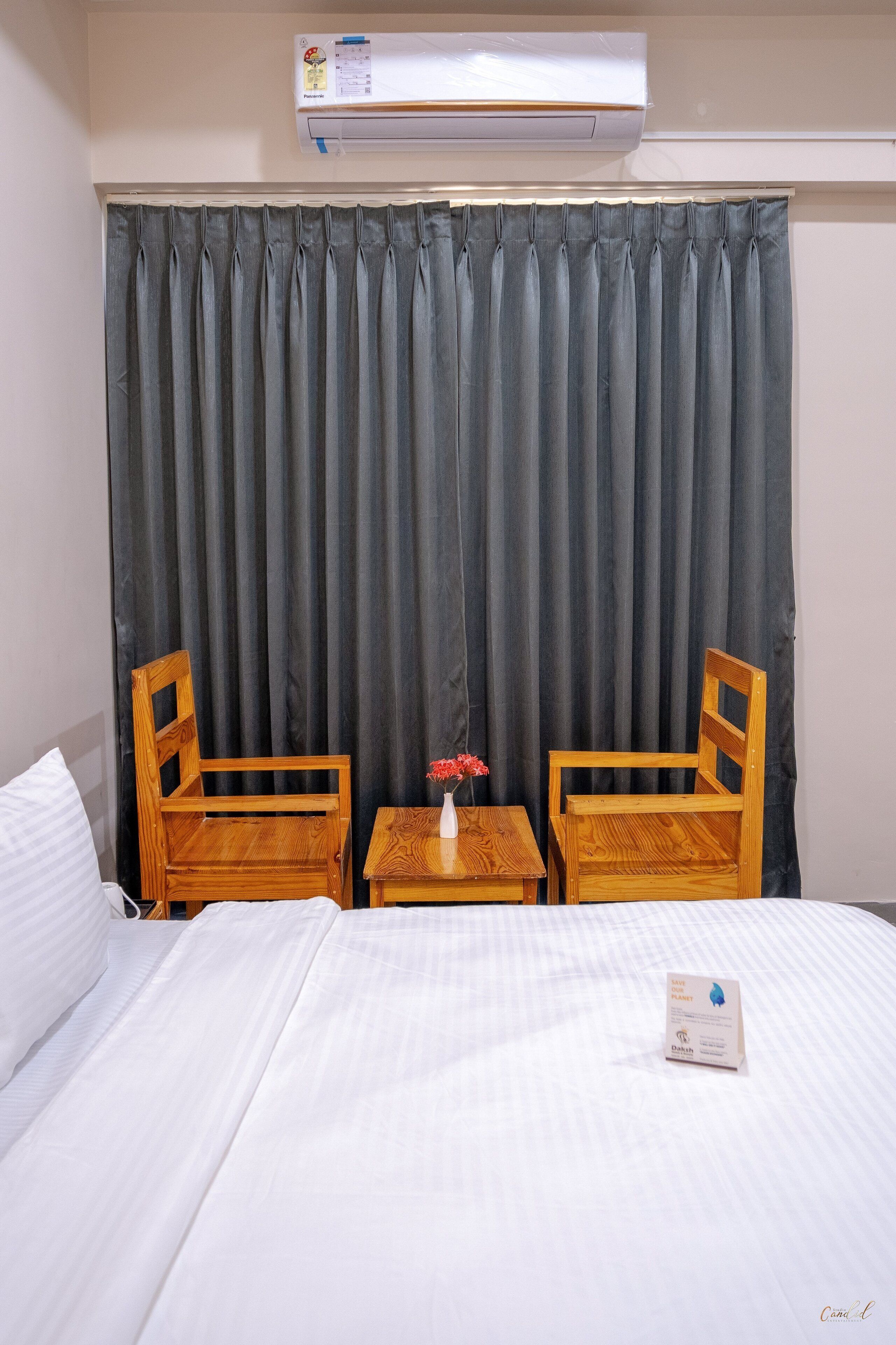 Habitación de hotel con cama, dos sillas de madera, una mesita con un florero y un aire acondicionado en la pared.