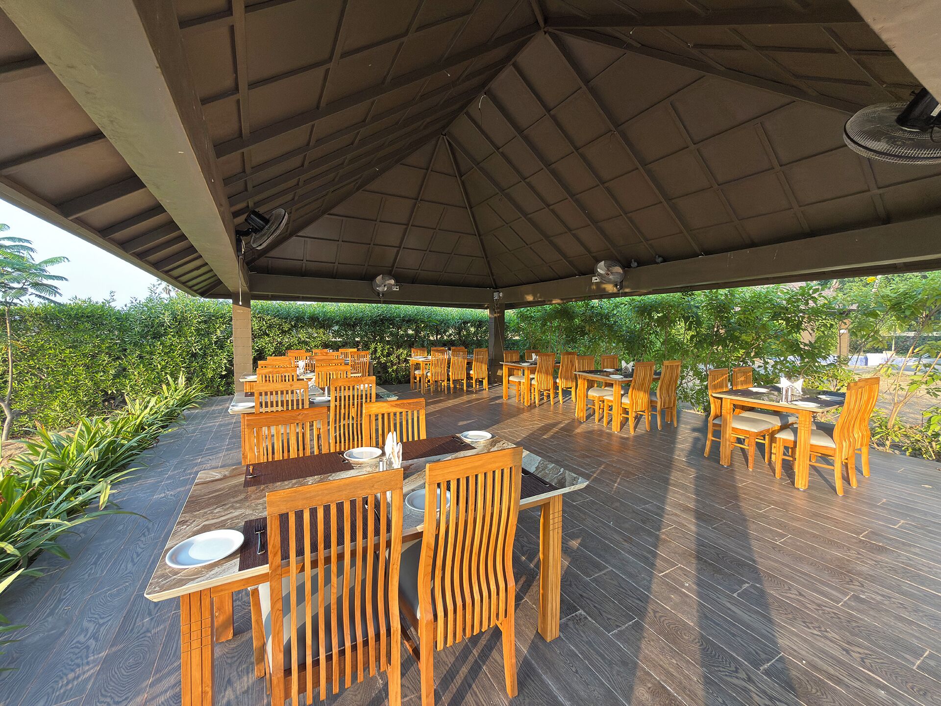 An outdoor dining area with multiple wooden tables and chairs, a covered roof, and a view of greenery.