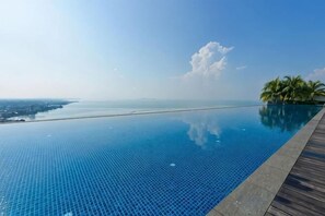 Una piscina infinita de color azul cristalino con una terraza de madera, palmeras y una vista distante de la costa.