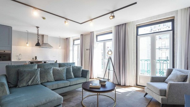 A modern living room with a sectional sofa, a round coffee table, and a grey armchair. There is a kitchen in the background with white cabinets and a hanging light fixture.