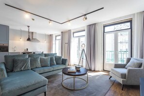 Una sala moderna con un sofá seccional, una mesa de centro redonda y un sillón gris. Se aprecia una cocina al fondo con armarios blancos y una lámpara colgante.