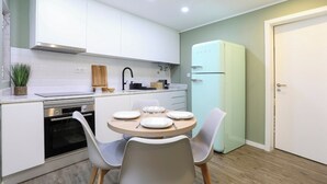 Een compacte keuken met witte kastjes, een ingebouwde oven, een spoelbak en een koelkast. Er staat een eettafel met stoelen en een potplant op het aanrecht.