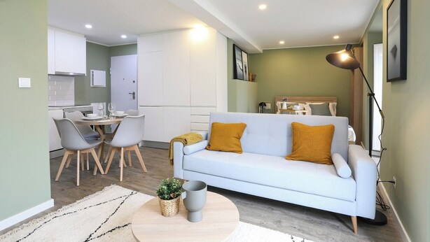 A modern living room with a light blue sofa, two mustard pillows, a coffee table with a plant and a cup, a dining area with a white table and chairs, and a kitchen in the background. - Amazing Studio @ Graça, Lisbon (Lisbon)