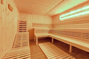 A sauna with wooden benches and benches, a sloped floor, and a wall-mounted light fixture. - GreenApartments (Chorzow)