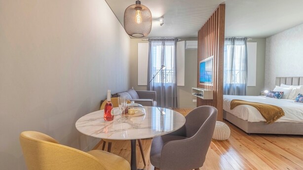 Een moderne hotelkamer met een ronde eettafel, twee stoelen, een bed met televisie en een groot raam met gordijnen.