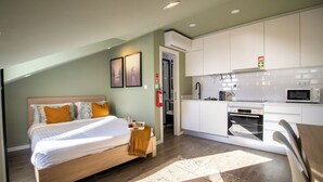 A bedroom with a bed, a bedside table, and a nightstand. There is a kitchenette with a microwave, a stove, and a sink. The room has a window and a door leading to the bathroom.