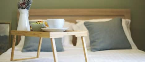 A neatly made bed with a wooden bedside table holding a white cup, a bowl, and a vase with dried flowers.