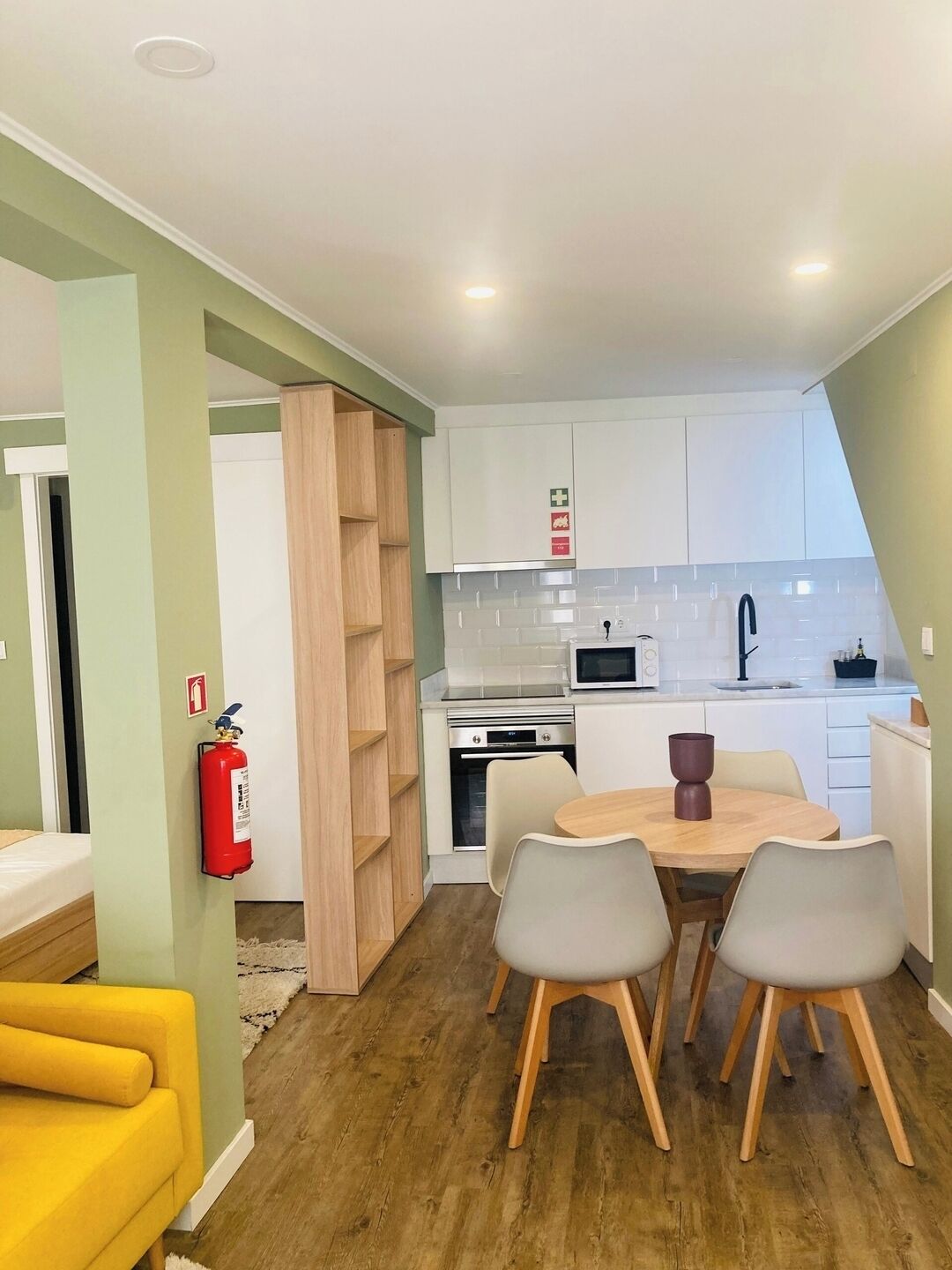A compact kitchen with white cabinets, a microwave, and a sink. There is a dining table with four chairs and a fire extinguisher mounted on the wall.