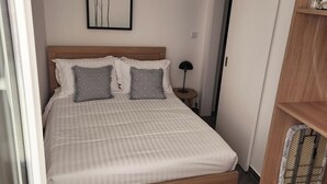 A neatly arranged bedroom with a bed, pillows, a bedside lamp, a framed picture on the wall, and a wooden shelf.