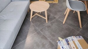 A modern living room with a grey sofa, a small round wooden table, and two grey chairs. A wooden partition with a crisscross design separates the living area from a kitchen with white cabinets and a built-in oven.