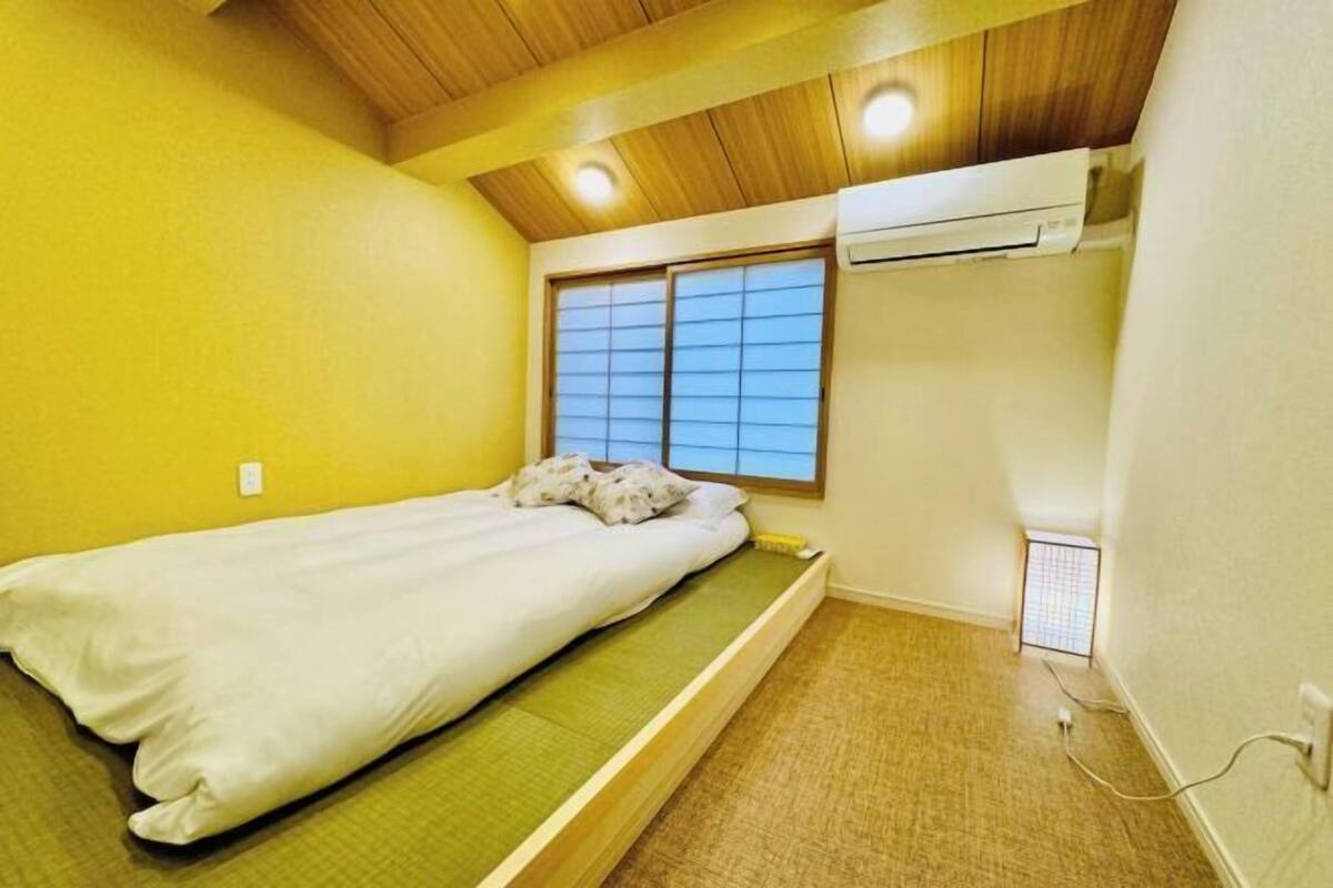 A small, single-bed room with a wooden ceiling, a window with blinds, and an air conditioning unit.