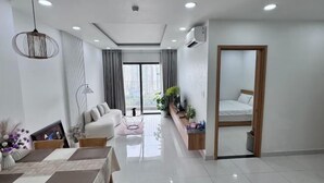 A modern living room with a sofa, dining table, and chairs. There is a small table with a vase of flowers. A bed is visible through an open door.