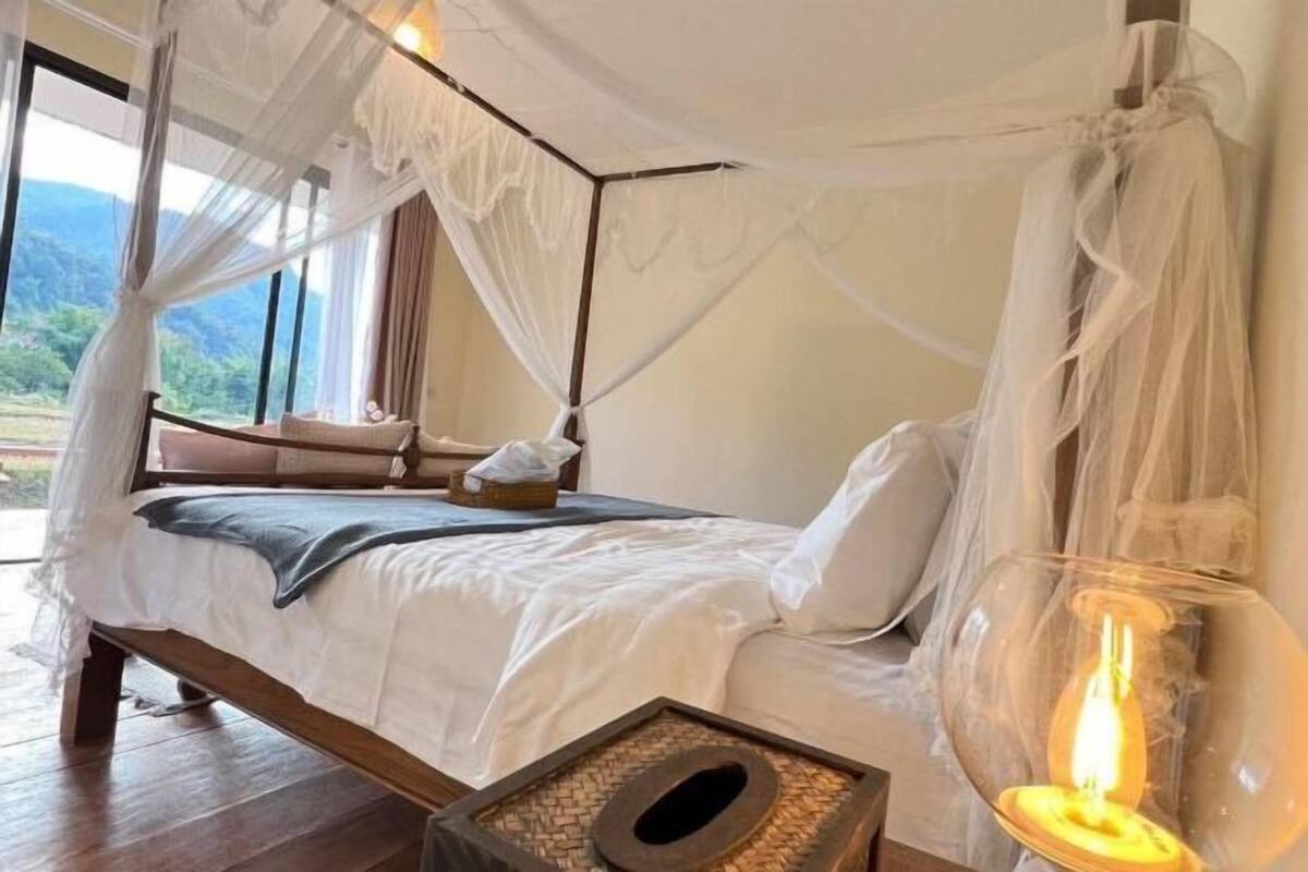 A four-poster bed with a canopy, a bedside table with a lamp, and a view of the outdoors.