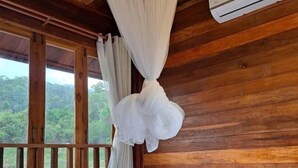 A wooden cabin room with a bed, a window with curtains, and a view of greenery outside. - Chan Rak Khao Villa (Bo Kluea)