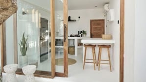 A modern kitchen with a white countertop, wooden stools, and a glass door leading to another room. - Villa Enda (Bali)