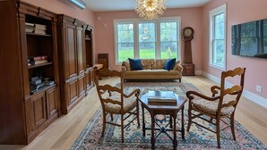 A spacious living room with a large wooden cabinet, a round table, and four chairs. There is a sofa with blue cushions, a chandelier, and a large window with a view of trees.