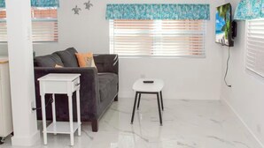 Ein Wohnzimmer mit einem grauen Sofa, einem weißen Beistelltisch und einem kleinen weißen Hocker. Zwei Fenster mit Jalousien, und ein Flachbildfernseher ist an der Wand montiert.
