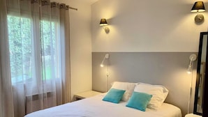 A neatly made bed with white linens and two blue pillows, a bedside table with a lamp, and a window with sheer curtains. - Mini villa 5 (Propriano)