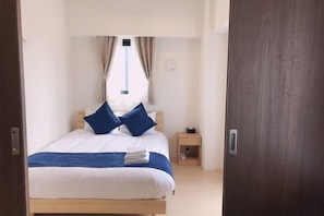 A neatly made bed with a blue and white bedspread, two blue pillows, and a white towel on the side. There is a wooden nightstand with a small lamp beside the bed. The room has a wooden door and a window with curtains.