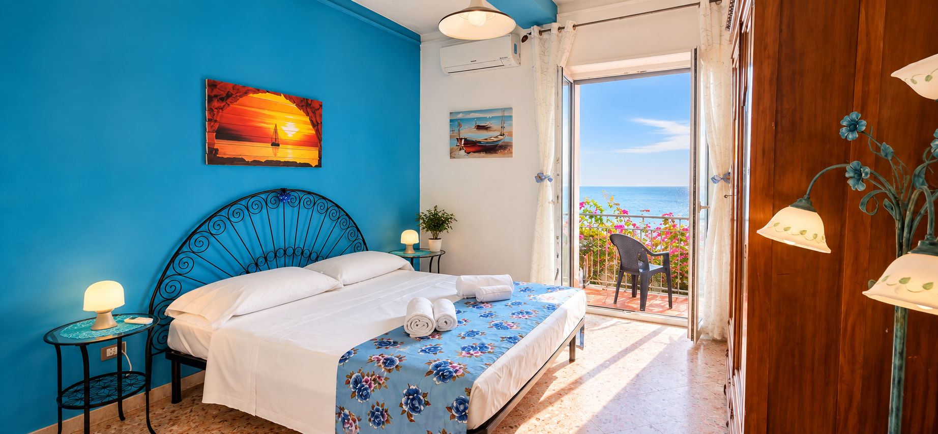 A bedroom with a bed, a nightstand, a lamp, and a view of the sea through an open door.