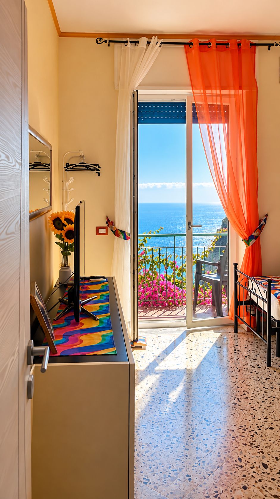 Een kamer met uitzicht op zee, een balkon met bloemen, een deur met spiegel en een kleurrijk vloerkleed.