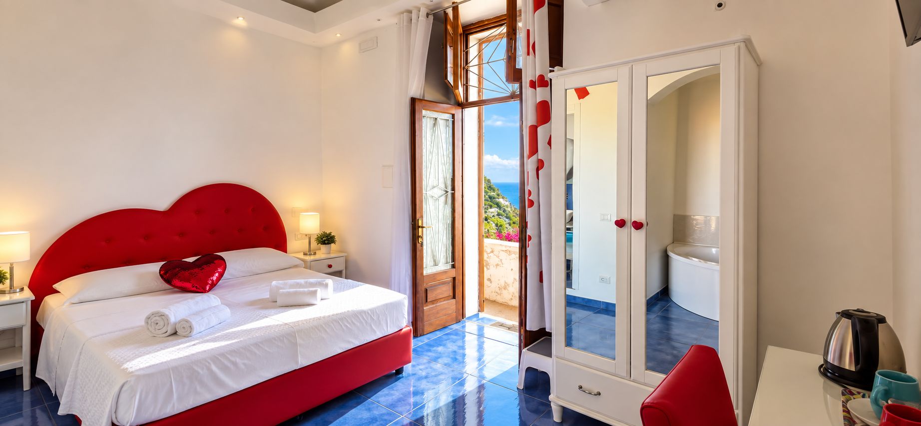 A hotel room with a red bed, white curtains, a view of the sea, a white wardrobe, and a coffee table with a kettle.