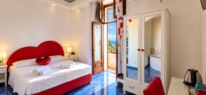 Een hotelkamer met een rode bed, witte gordijnen, uitzicht op zee, een witte kast en een bijzettafeltje met een waterkoker.