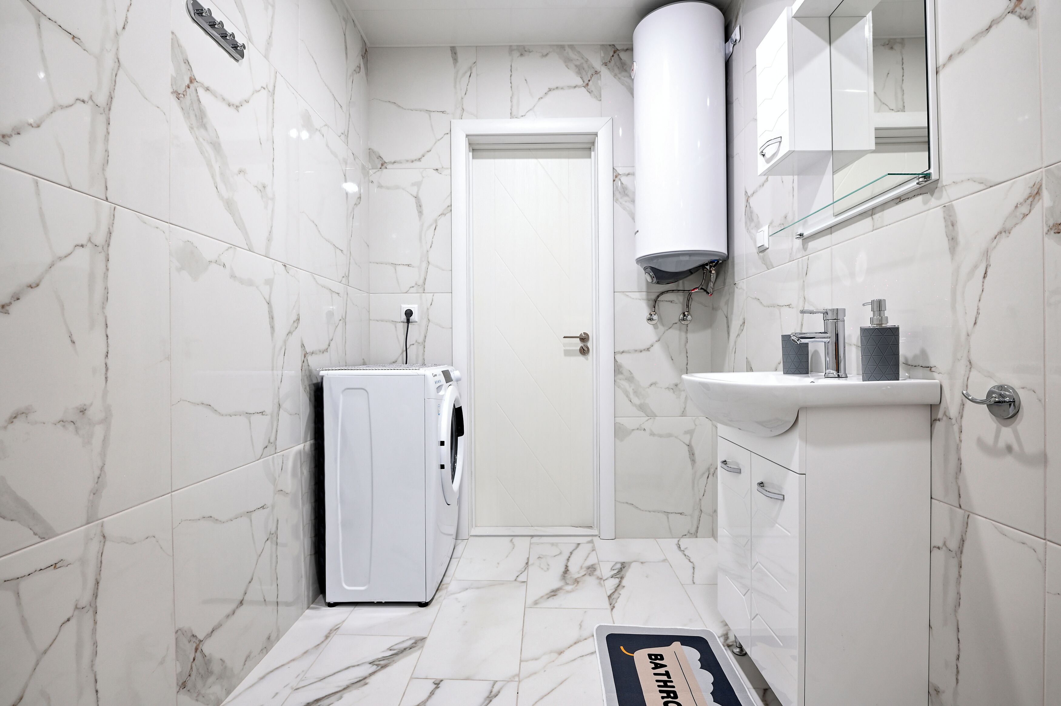 Une salle de bain avec des carreaux de marbre blanc, un lavabo blanc, un miroir, un porte-serviettes et une porte.