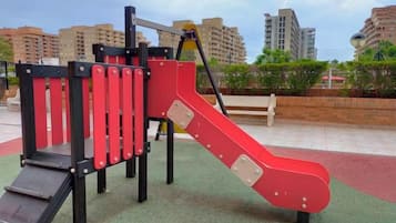 Un parque infantil con tobogán y estructuras para escalar, rodeado de una zona pavimentada y edificios al fondo.