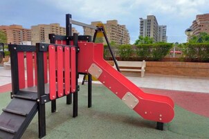 A playground with a slide and climbing structures, surrounded by a paved area and buildings in the background.