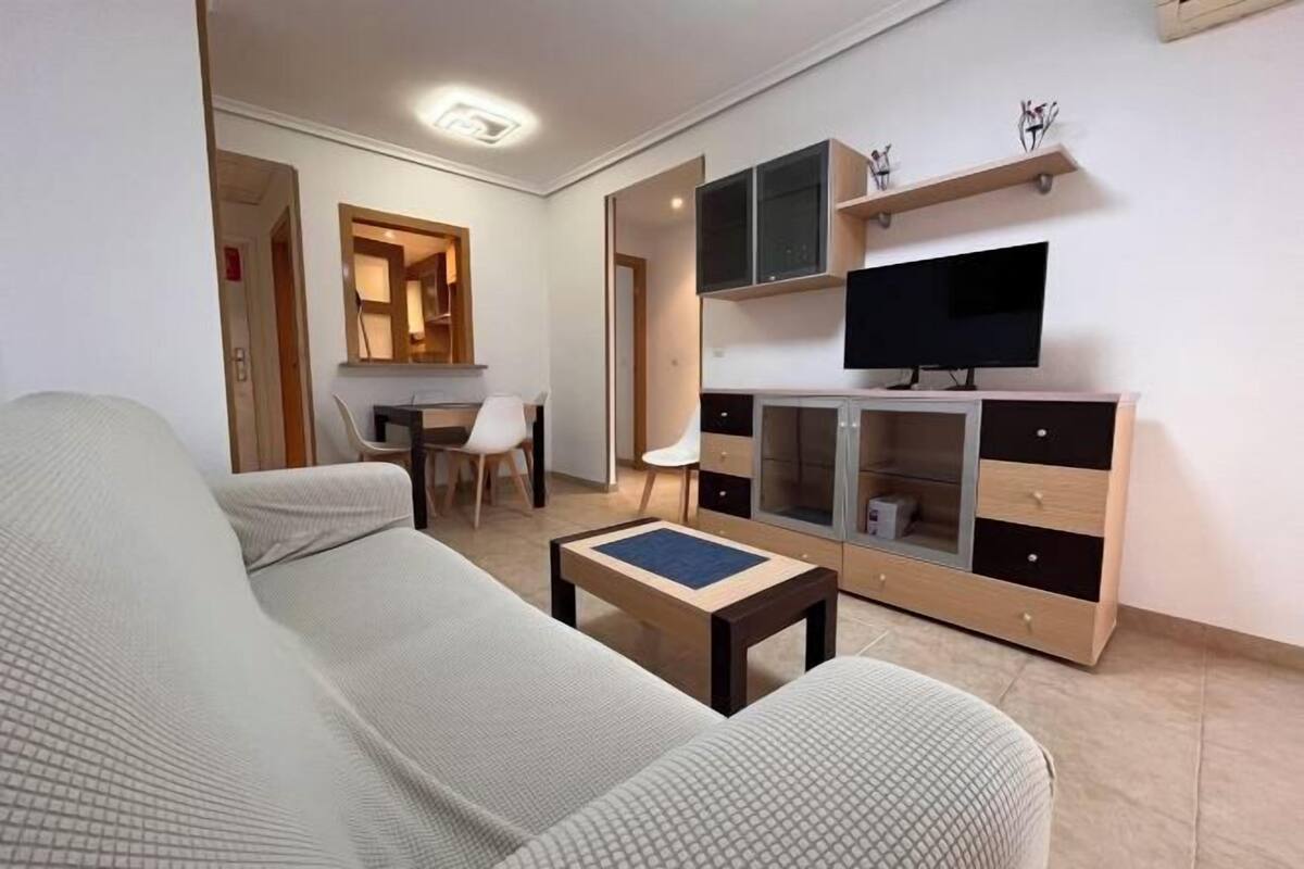 Una sala moderna con un sofá, una mesa de centro, un televisor sobre un mueble, y un comedor con una silla y una mesa.