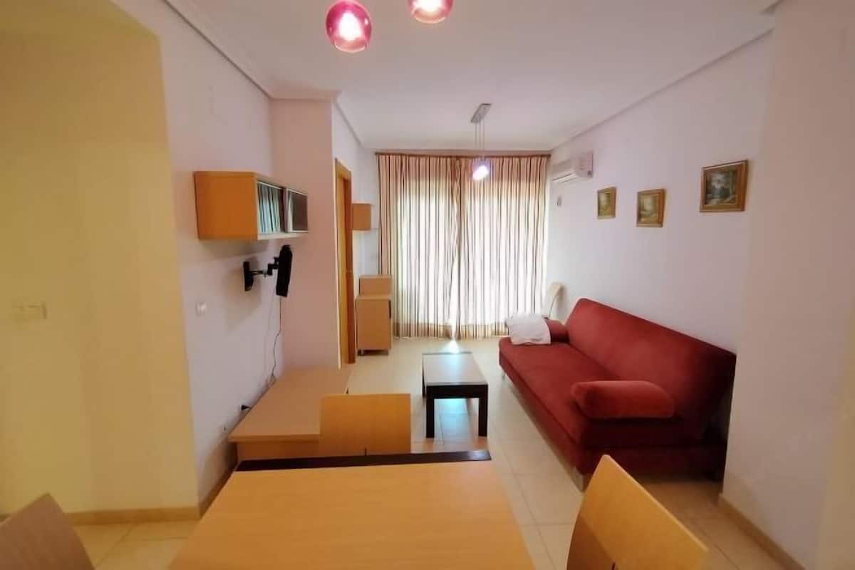 Una sala de estar con un sofá rojo, una mesa de centro de madera, un televisor montado en la pared y un ventanal con cortinas.