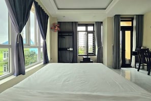 Une chambre d’hôtel avec un grand lit, un bureau et une chaise. Il y a une fenêtre avec des rideaux, et on peut voir un ventilateur au plafond.