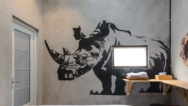 A room with a large black and white rhinoceros mural, a bed, a desk with a computer, and a small trash bin. - Exotic Inn Semarang, by INNCA COLLECTION (Semarang)