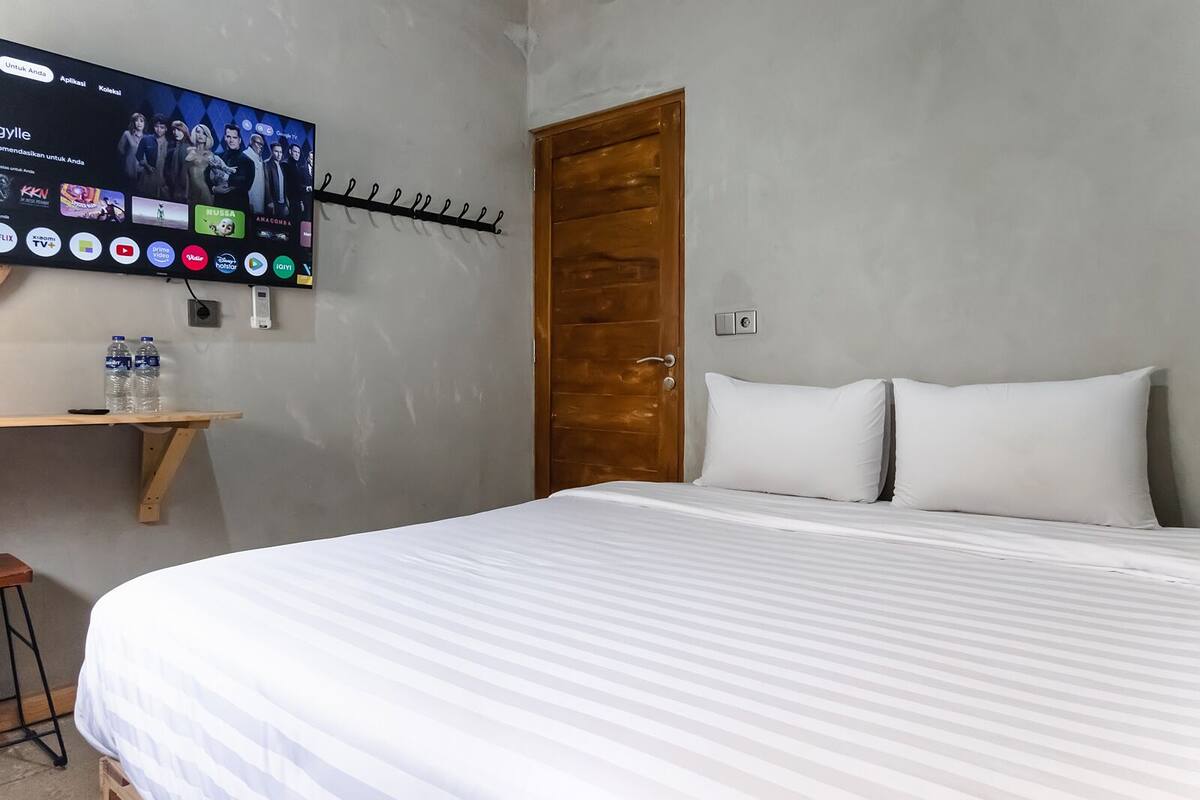 A hotel room with a bed, a TV mounted on the wall, a wooden door, and a small wooden shelf with water bottles.