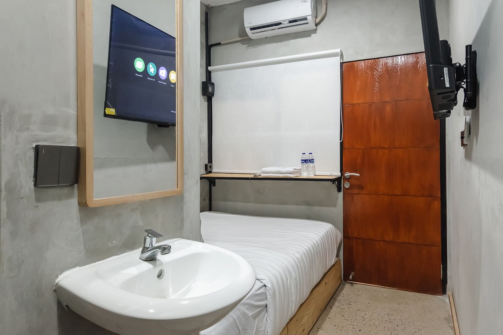 A compact hotel room with a single bed, a white sink, a mirror, a mounted TV, and a wooden door.