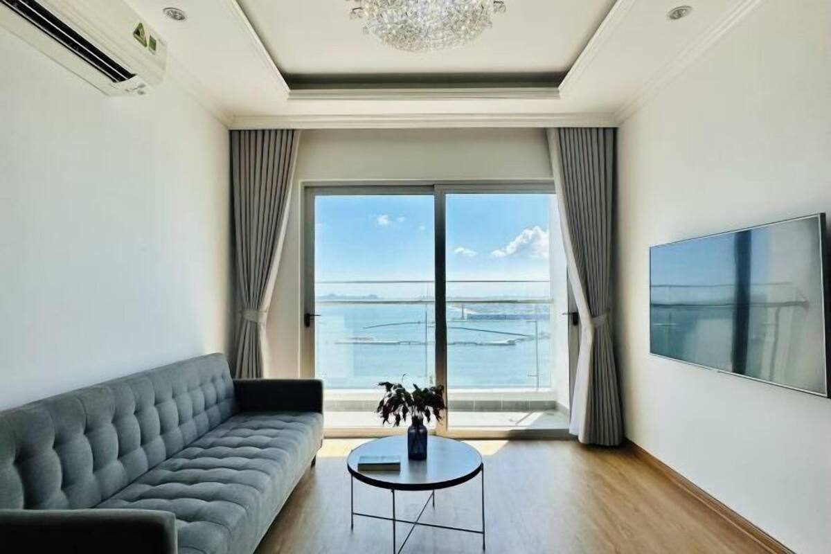 A modern living room with a sofa, a round coffee table, a TV, and a large window with a view of the sea.