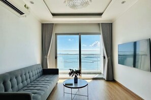 A modern living room with a sofa, a round coffee table, a TV, and a large window with a view of the sea. - Cozy Homestay 2 BR sea view 29th flr Halong center. (Ha Long)