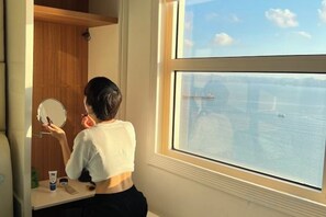 A person in a room with a window overlooking a body of water, a mirror, and a small table with items on it. - Cozy Homestay 2 BR sea view 29th flr Halong center. (Ha Long)