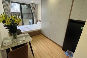 A compact hotel room with a bed, a small table with a vase of flowers, a chair, a refrigerator, and a window with curtains.