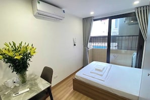A hotel room with a bed, a table with a vase of flowers, an air conditioning unit, and a view of a building and pool.