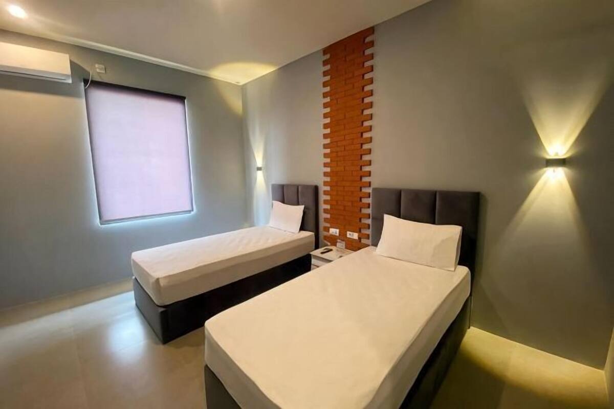 Habitación de hotel con dos camas, una ventana con persianas, un muro decorativo de ladrillo e iluminación empotrada.