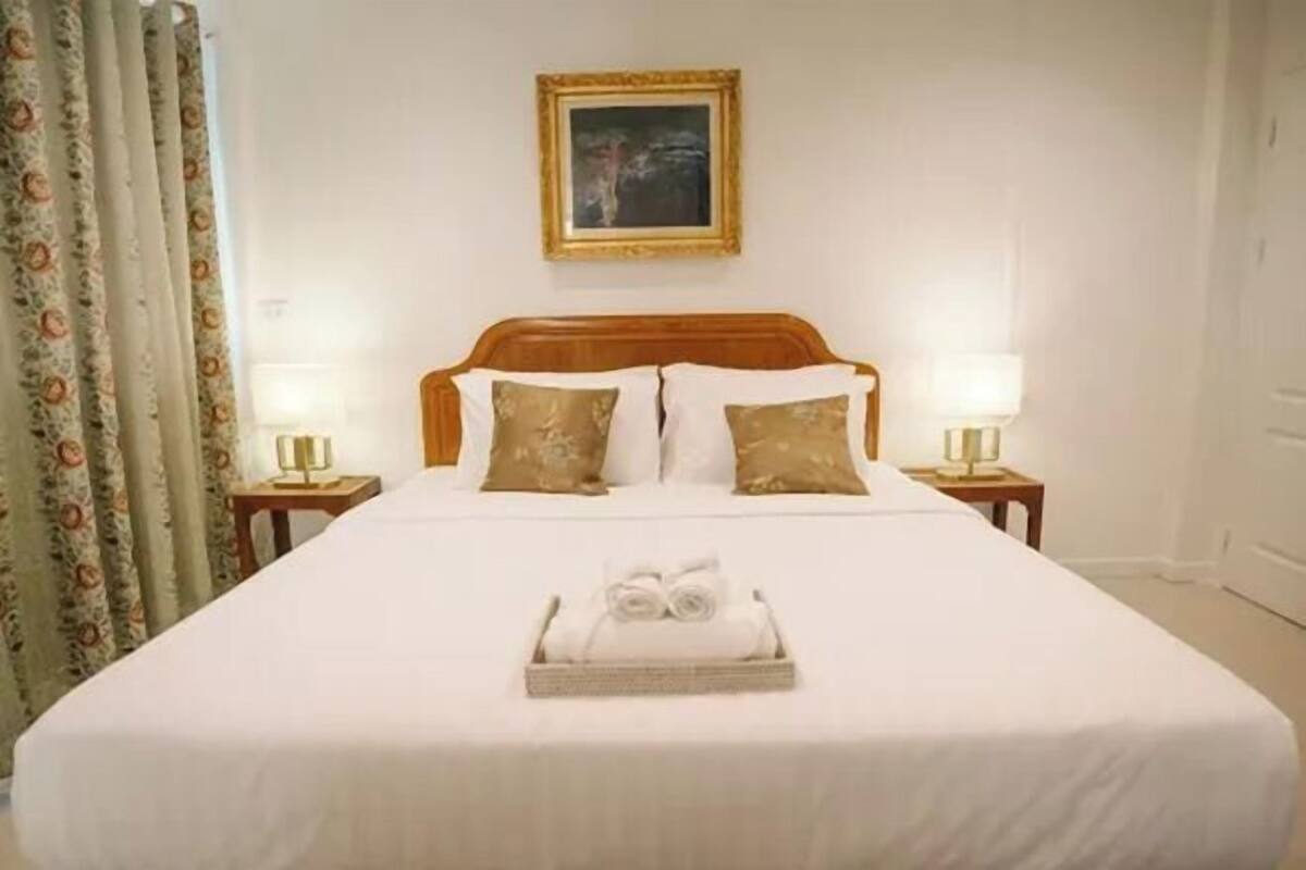 A neatly made bed with a white bedspread and two pillows, a framed picture on the wall, and a tray with folded towels on the bed.