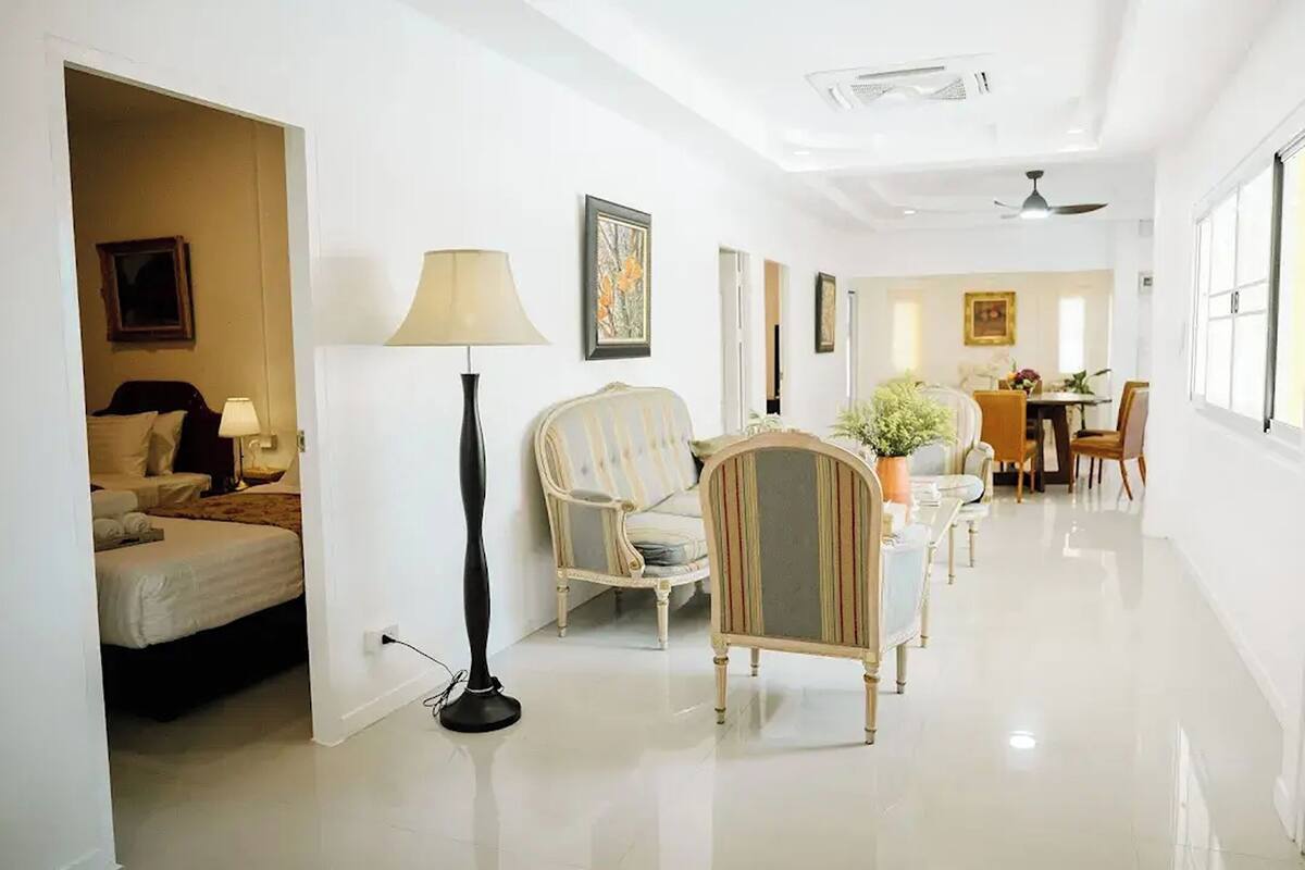 A hotel room with a bed, a standing lamp, a striped armchair, and a dining area with a table and chairs.